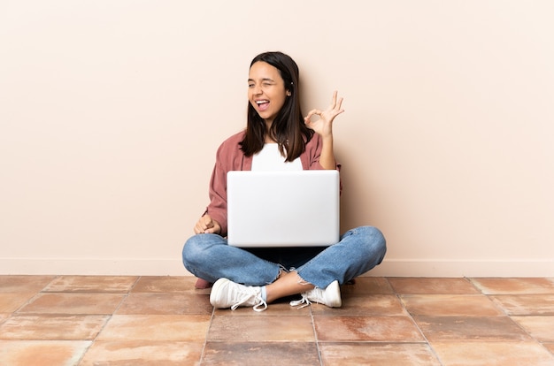 Young mixed race woman with a laptop sitting on the floor showing ok sign with fingers