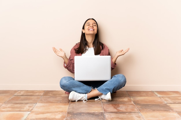 Young mixed race woman with a laptop sitting on the floor smiling a lot