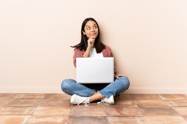 Young mixed race woman with a laptop sitting on the floor thinking an idea while looking up