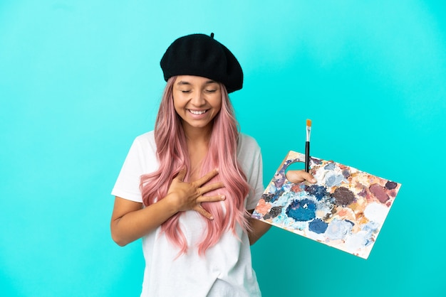 Photo young mixed race woman with pink hair holding a palette isolated on blue background smiling a lot