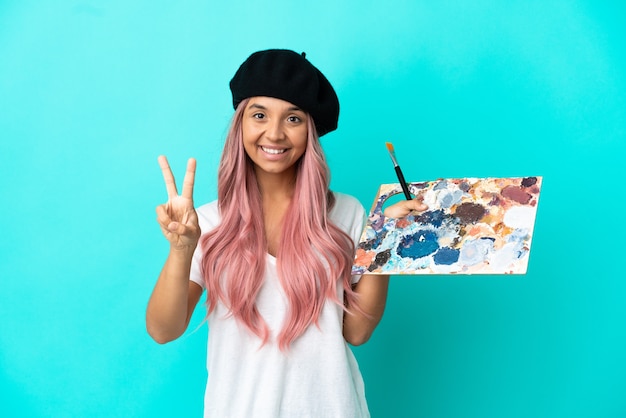 Photo young mixed race woman with pink hair holding a palette isolated on blue background smiling and showing victory sign