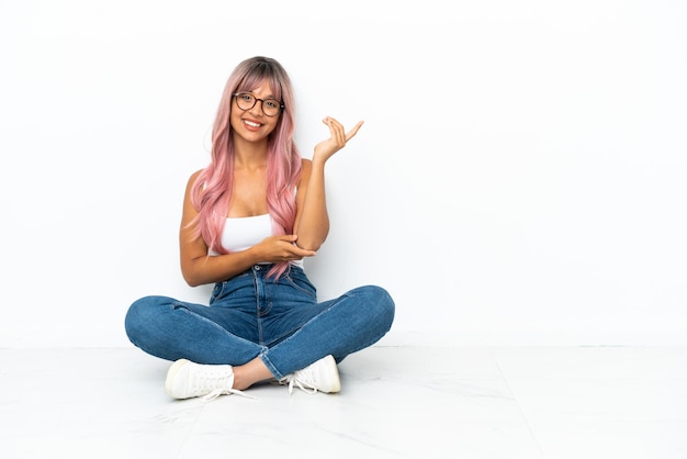 Young mixed race woman with pink hair sitting on the floor isolated on white background extending hands to the side for inviting to come