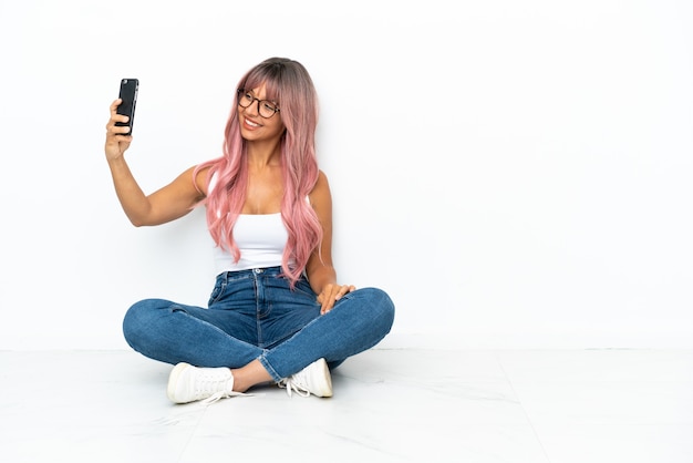 Young mixed race woman with pink hair sitting on the floor isolated on white background making a selfie