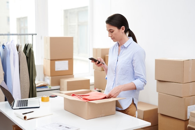 Young mobile female with smartphone taking photo of folded pullover in open box before packing and sending it to client