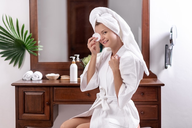 Young model in a bathrobe and a towel on her head sitting on a chair near the table 