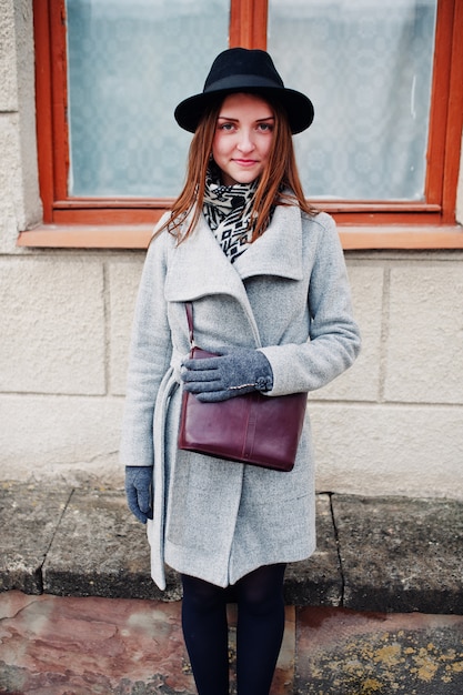 Young model girl in a gray coat and black hat with leather handbag on shoulders against window of old house at street of city.