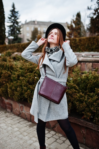 Young model girl in a gray coat and black hat with leather handbag on shoulders posed near bushes at street of city.