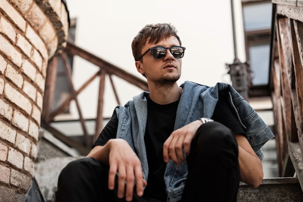 Young modern hipster man in a fashionable denim jacket in black pants in stylish dark sunglasses relax sitting on a vintage staircase near brick wall in the city. Cool guy enjoys a weekend outdoors.