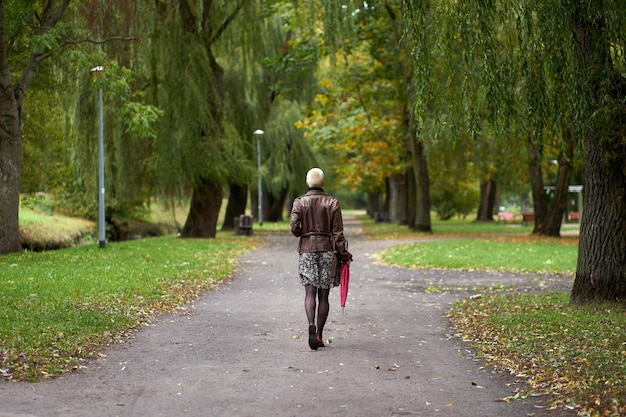 Young modern short haired blonde woman walking in the autumn park with red umbrella View from back