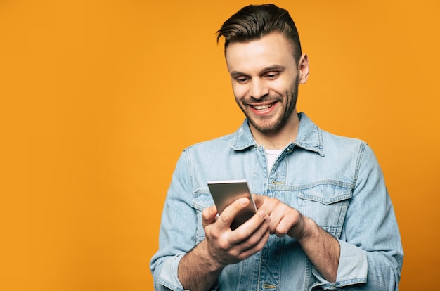 Young modern stylish man in denim is using smartphone in his hands while standing over yellow background