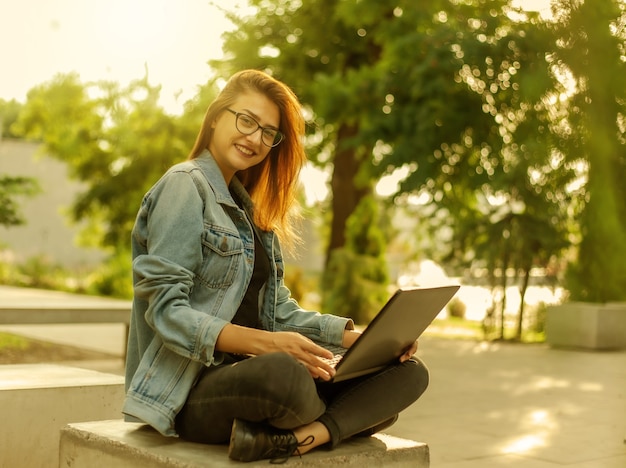 Young modern woman blogger in a denim jacket and glasses holds a laptop in his hands in the park. Online learning, blogging