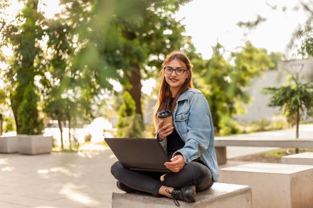 Young modern woman student in a denim jacket sitting at park and uses laptop with coffee cup on hand. Distance learning. Modern youth concept.