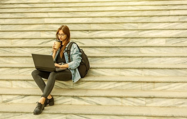 Young modern woman student in a denim jacket sitting on the stairs with laptop. Distance learning. Modern youth concept.