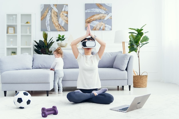 Young mom meditating in lotus yoga position using ar glasses while her daughter watches cartoons on