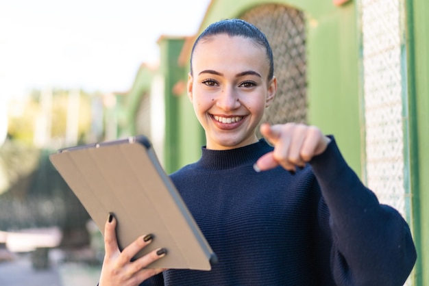 Young moroccan girl holding a tablet at outdoors points finger at you with a confident expression