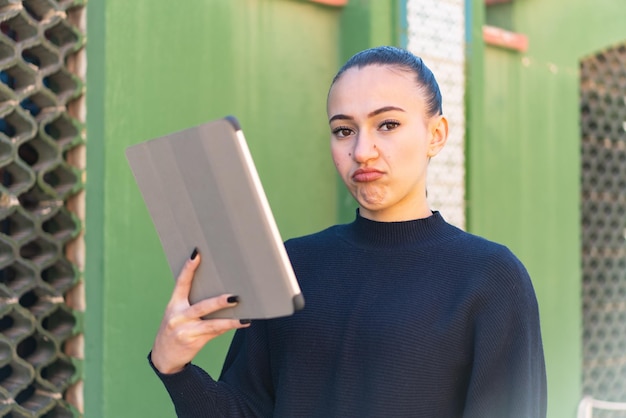 Young moroccan girl holding a tablet at outdoors with sad expression