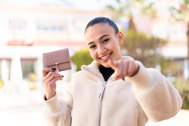 Young moroccan girl holding a wallet at outdoors points finger at you with a confident expression