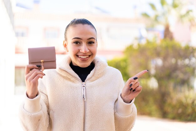 Young moroccan girl holding a wallet at outdoors surprised and pointing finger to the side