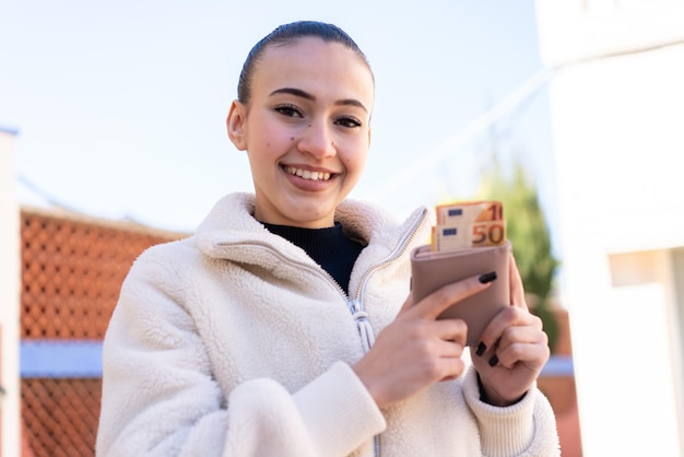 Young moroccan girl at outdoors holding wallet with money with happy expression