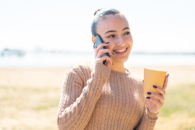 Young moroccan girl at outdoors using mobile phone and holding a coffee with happy expression