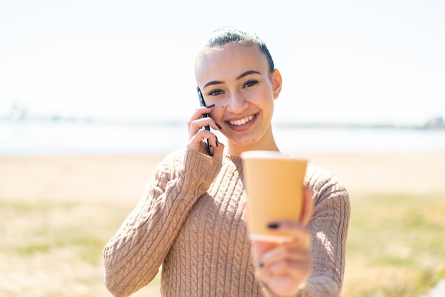 Young moroccan girl at outdoors using mobile phone and holding a coffee with happy expression