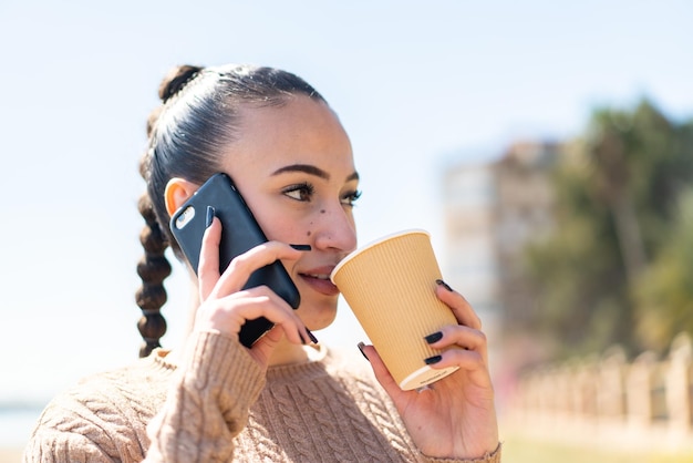 Young moroccan girl at outdoors using mobile phone and holding a coffee with happy expression