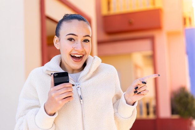 Young moroccan girl using mobile phone at outdoors surprised and pointing finger to the side