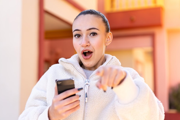 Young moroccan girl using mobile phone at outdoors surprised and pointing front