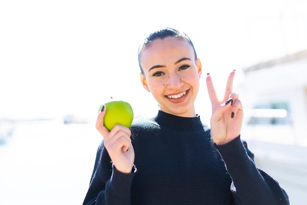 Young moroccan girl with an apple at outdoors smiling and showing victory sign