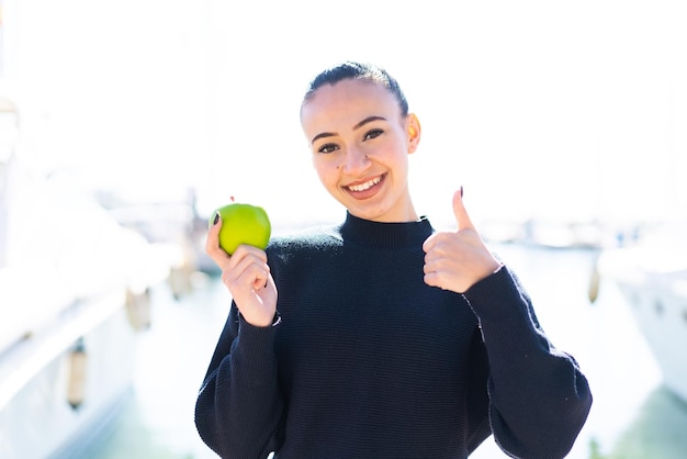 Young moroccan girl with an apple at outdoors with thumbs up because something good has happened