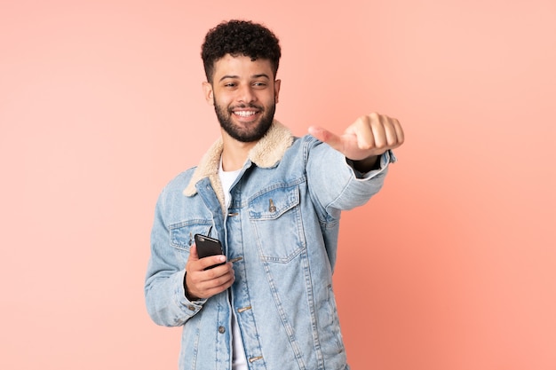 Young Moroccan man using mobile phone isolated on pink wall giving a thumbs up gesture