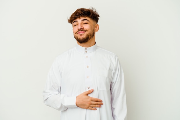 Young Moroccan man wearing a typical arab clothes isolated on white touches tummy, smiles gently, eating and satisfaction concept.