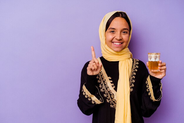 Young Moroccan woman holding a glass of tea isolated on purple background showing number one with finger.