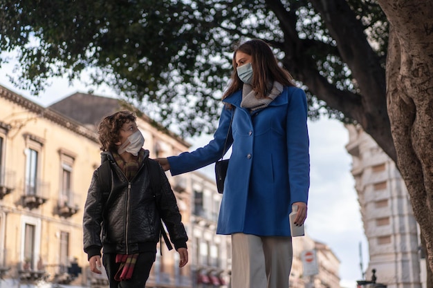 A young mother accompanies her son to school on the street. Both wear a protective face mask to prevent the spread of the coronavirus. New normal back to school concept.