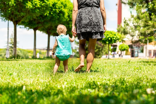 Young mother and barefoot daughter holding hands walk barefoot on a green lawn