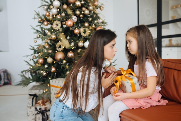 Young mother calming down her sad infant daughter sitting on the Christmas tree background