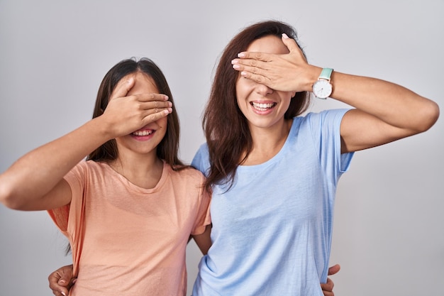 Young mother and daughter standing over white background smiling and laughing with hand on face covering eyes for surprise blind concept