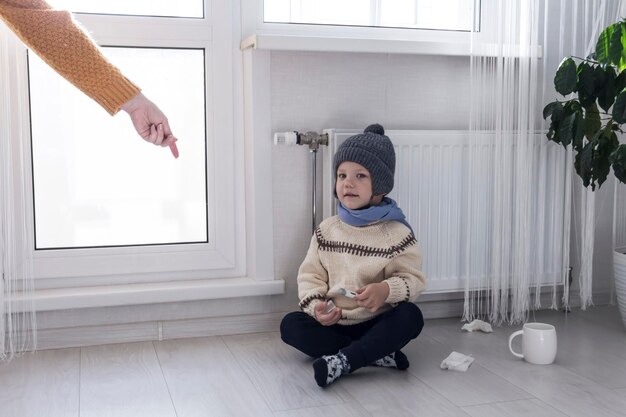 A young mother gives medicine to her little son who is warmly dressed and sitting on the floor