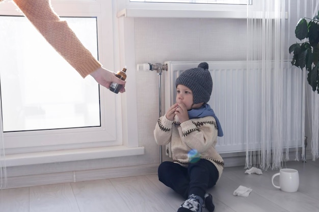 A young mother gives medicine to her little son who is warmly dressed and sitting on the floor