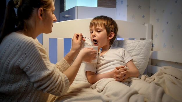 Young mother giving medicines and syrup from spoon to her sick little son lying in bed