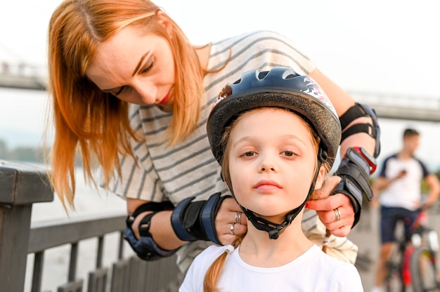 Young mother helping her little daughter to put a bicycle helmet on. Preschool girl having active walk with her mother