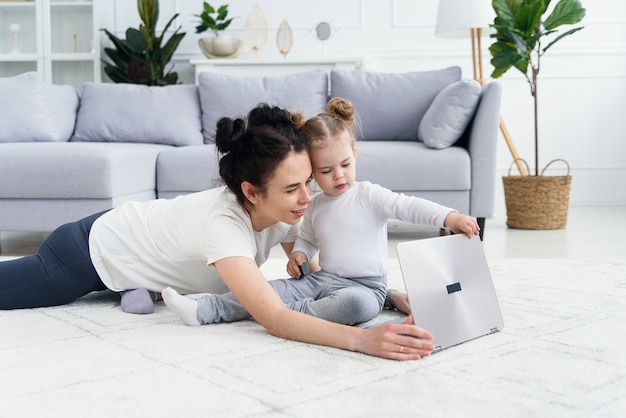 Young mother and her adorable daughter are using laptop while lying on the floor.