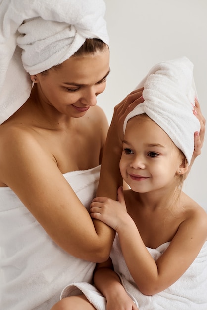 young mother and her little daughter in white towels in the bedroom in the morning. Happy family at home.