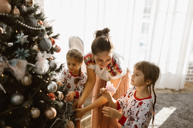 Young mother and her little daughters decorate a New Year's tree in the light cozy room