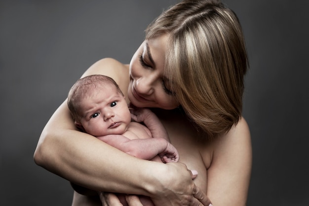 A young mother holds a newborn baby 1 week old in her arms. Love and tenderness in breastfeeding. Close-up. Gray background, toning.