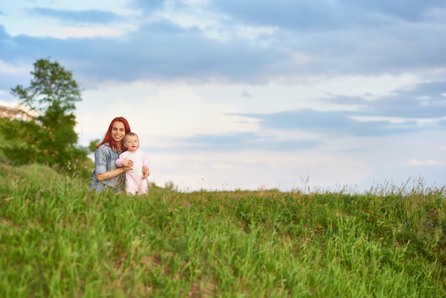 Young mother hugging cute little daughter sitting on grass in field.