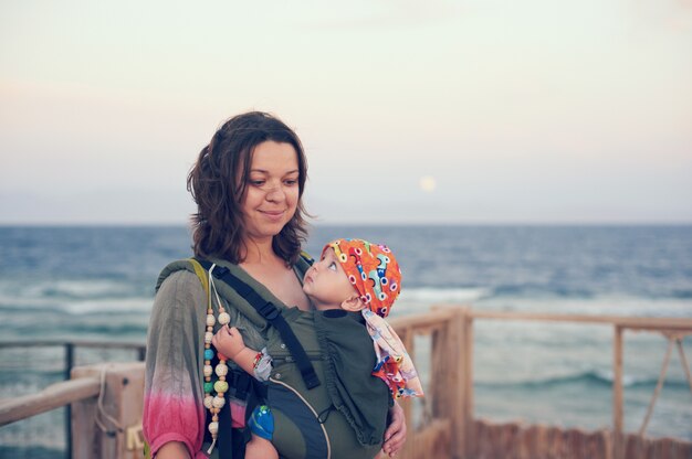 A young mother is on the beach with her baby in a sling