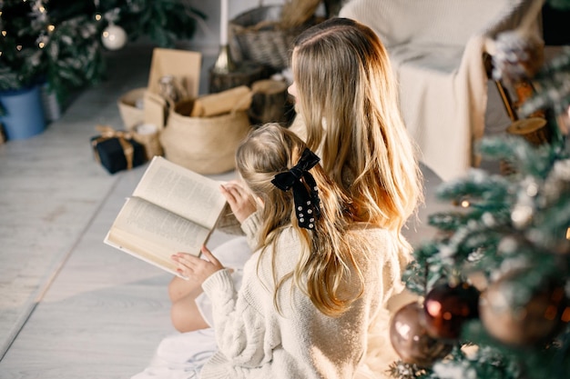 Young mother and little girl sitting christmas tree at home and reading a book