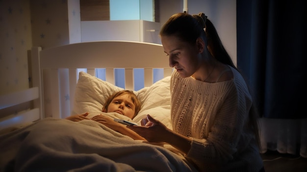 Young mother looking for doctor phone number on smartphone to call him to her sick son lying in bed