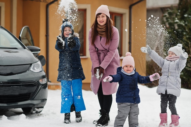 Young mother play with kids against electric car in the yard of her house at winter